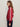 A woman with long brown hair, glasses, and hoop earrings stands in profile against a plain light background. She wears the Sanctuary Clothing satin track jacket crimson with white sleeve stripes and dark pants.