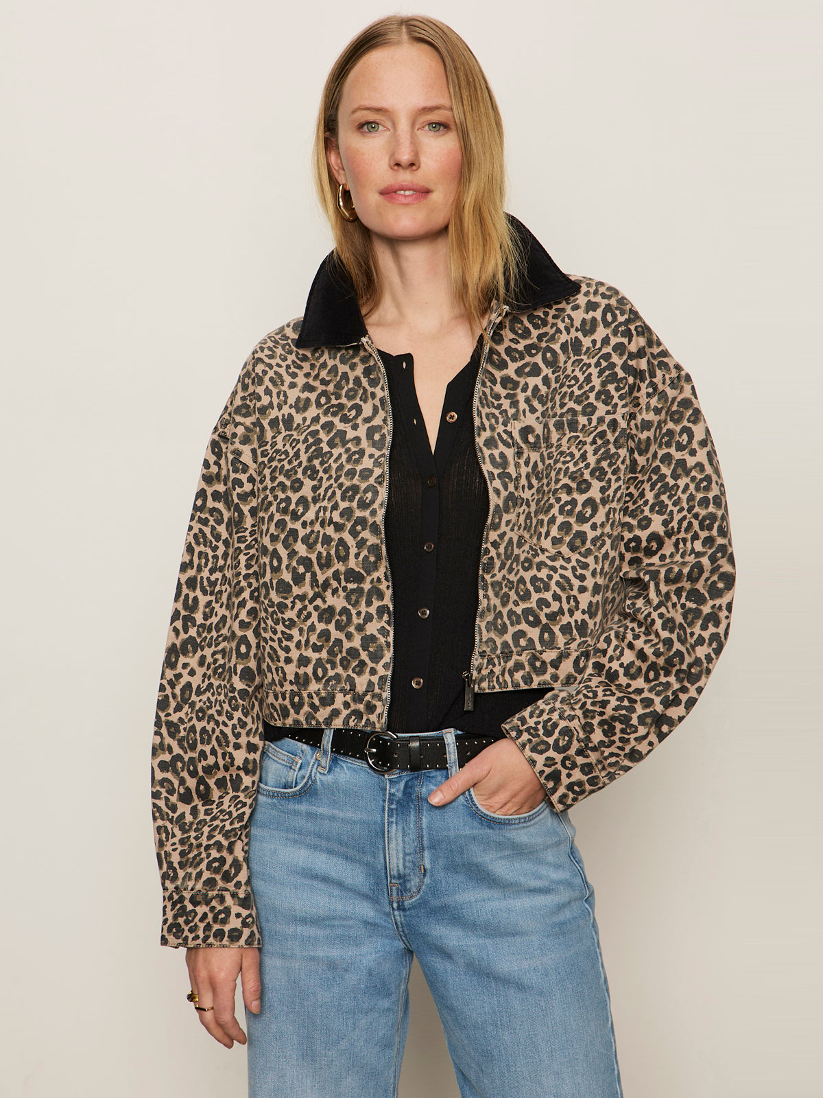 A woman with straight blonde hair and a relaxed expression models the Sanctuary Clothing cropped barn jacket french kitty—a leopard print jacket with a black collar—over a black top and light blue jeans against a plain background.