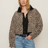A woman with straight blonde hair and a relaxed expression models the Sanctuary Clothing cropped barn jacket french kitty—a leopard print jacket with a black collar—over a black top and light blue jeans against a plain background.