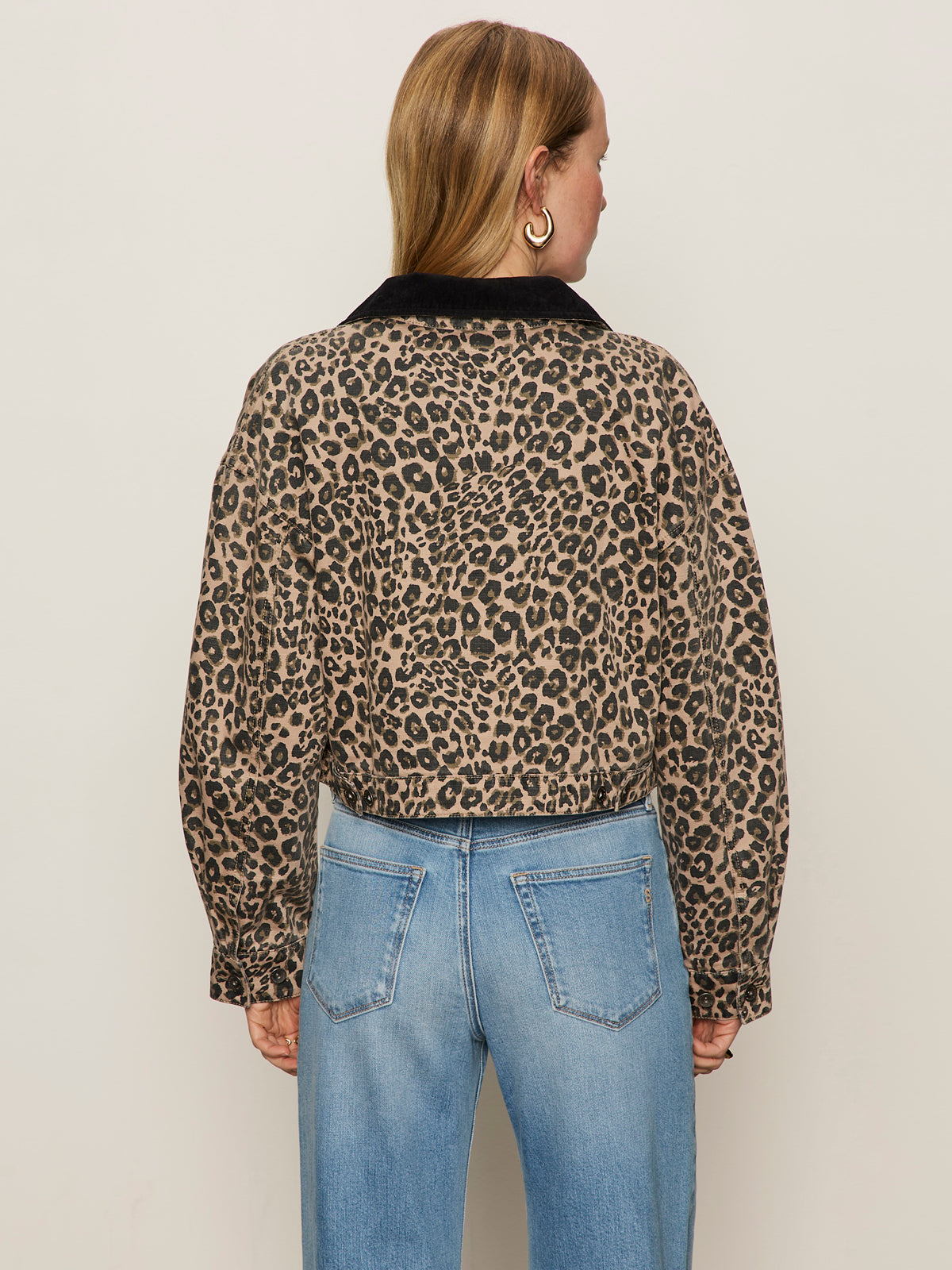 A person with light brown hair, wearing large hoop earrings and light blue jeans, is seen from behind in the Sanctuary Clothing cropped barn jacket french kitty, featuring a leopard print with black collar, against a plain light background.