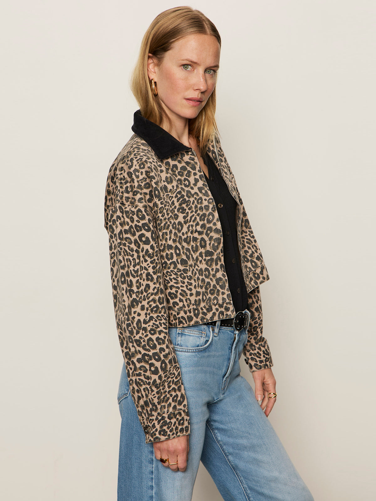 A woman with straight blonde hair poses confidently in the Sanctuary Clothing cropped barn jacket french kitty—a leopard-print piece with a black collar—paired with light blue jeans, one hand in her pocket against a plain background.