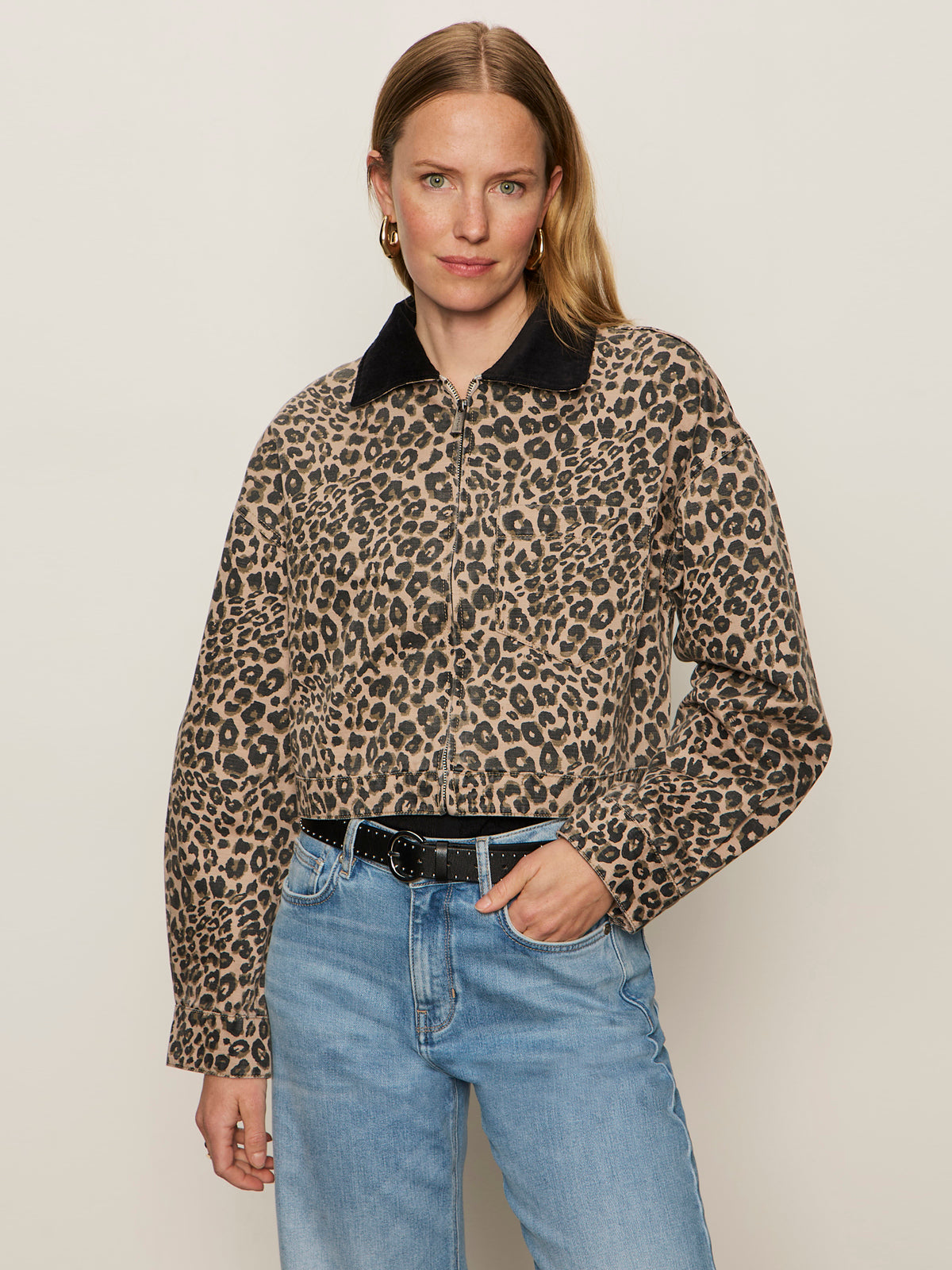 A woman with long, light brown hair models the Sanctuary Clothing cropped barn jacket french kitty—leopard print with a black collar—paired with light blue jeans, a black belt, and gold hoop earrings against an off-white background.