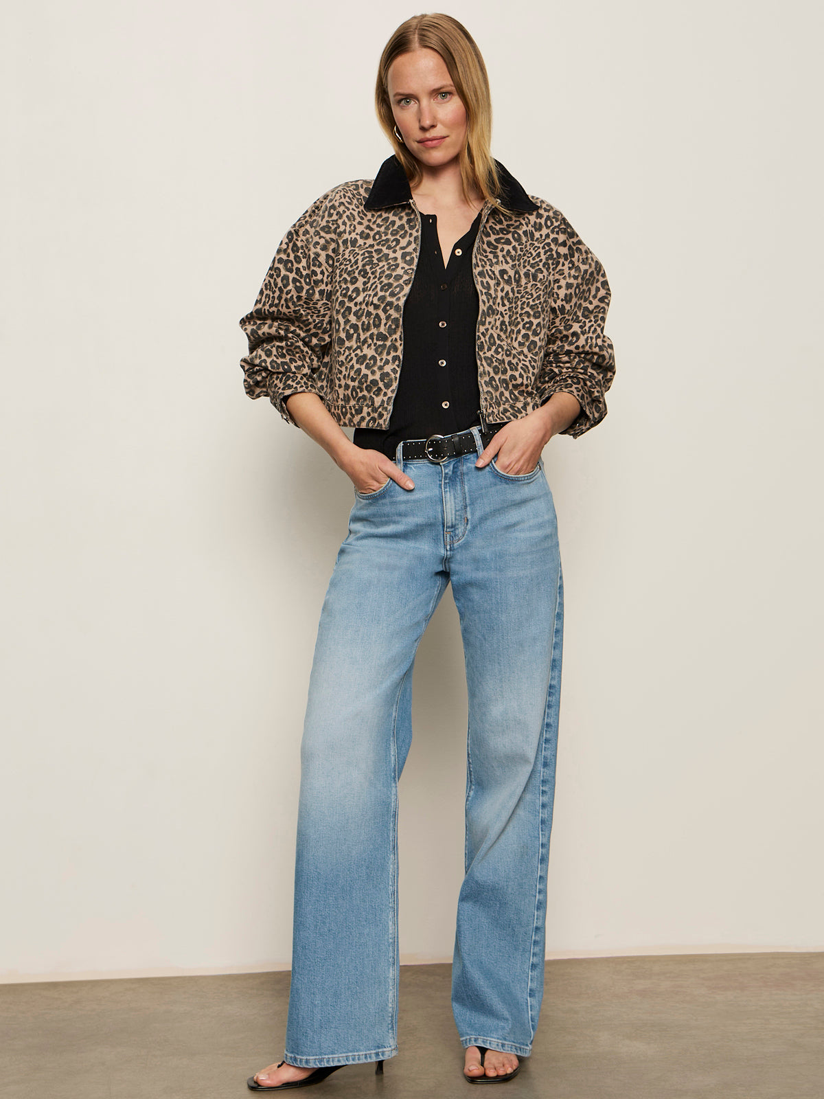A woman stands against a plain wall wearing the Sanctuary Clothing cropped barn jacket french kitty over a black top, light blue high-waisted wide-leg jeans, and black open-toe heels, one hand in her pocket.