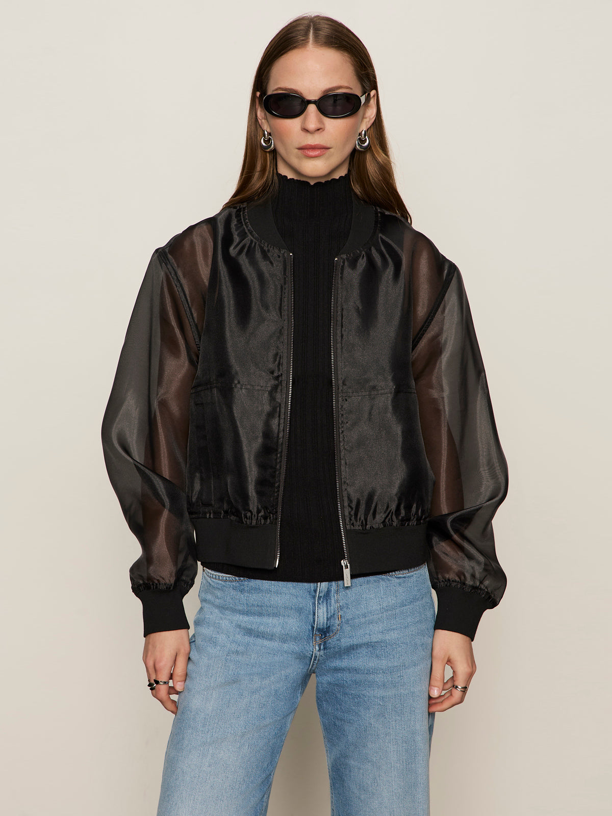 A woman with long brown hair wears Sanctuary Clothing's shane organza bomber black over a black turtleneck, paired with black oval sunglasses, silver hoop earrings, and light blue jeans against a plain light background.
