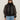 A woman with long brown hair wears Sanctuary Clothing's shane organza bomber black over a black turtleneck, paired with black oval sunglasses, silver hoop earrings, and light blue jeans against a plain light background.