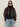 A woman with long brown hair wears Sanctuary Clothing's shane organza bomber black over a black turtleneck, paired with black oval sunglasses, silver hoop earrings, and light blue jeans against a plain light background.