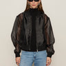 A woman with long brown hair wears Sanctuary Clothing's shane organza bomber black over a black turtleneck, paired with black oval sunglasses, silver hoop earrings, and light blue jeans against a plain light background.
