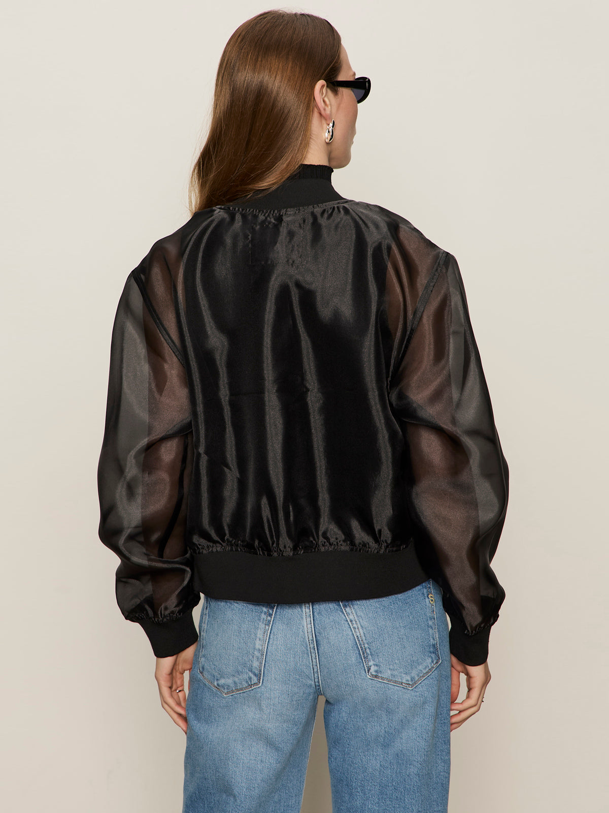 A person with long brown hair wears sunglasses and the Sanctuary Clothing shane organza bomber black jacket with light blue jeans, standing with their back to the camera against a plain light background.