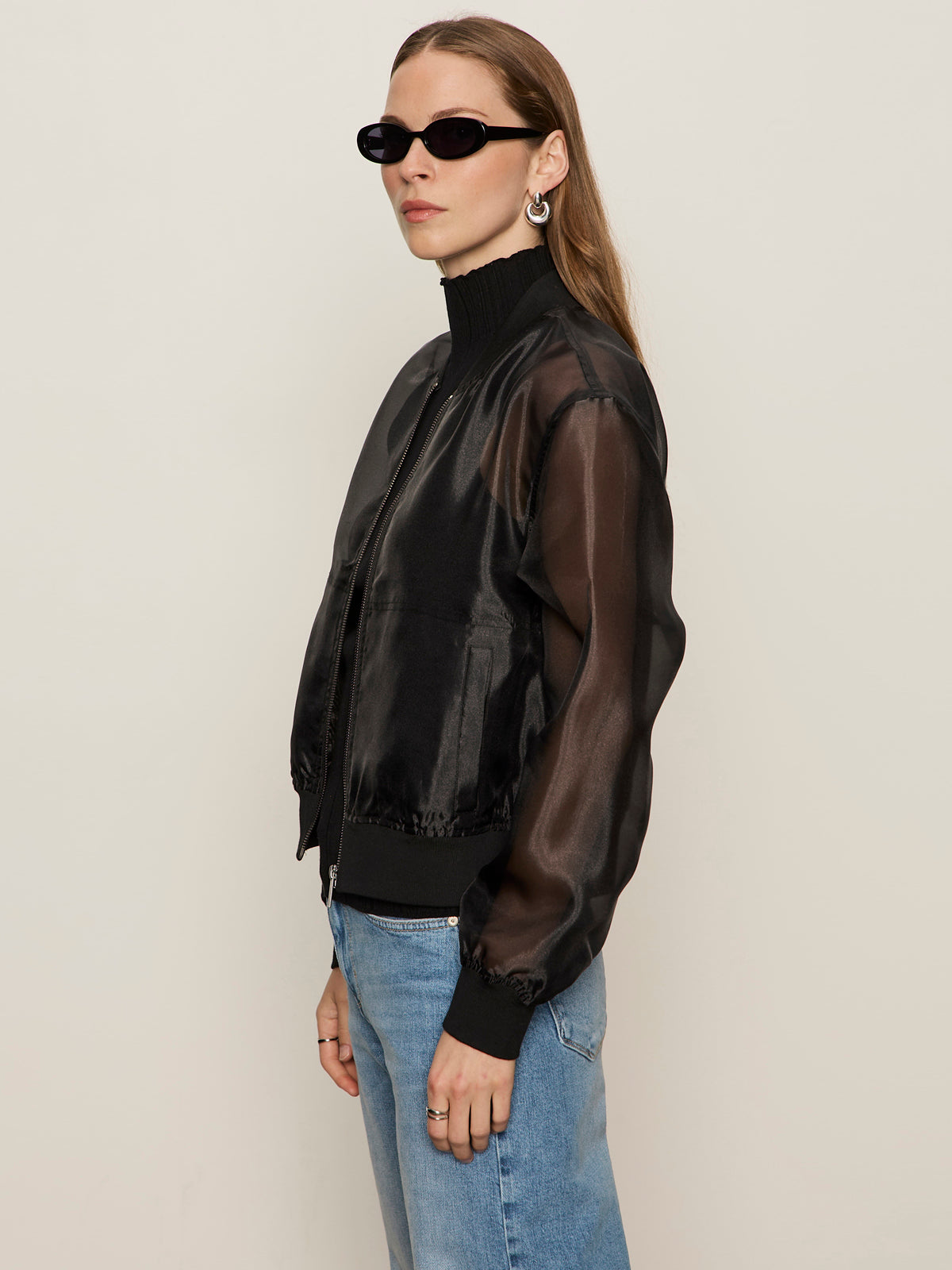 A woman with long brown hair wears Sanctuary Clothing's Shane organza bomber in black, black oval sunglasses, silver hoop earrings, and light blue jeans, standing against a plain beige background.