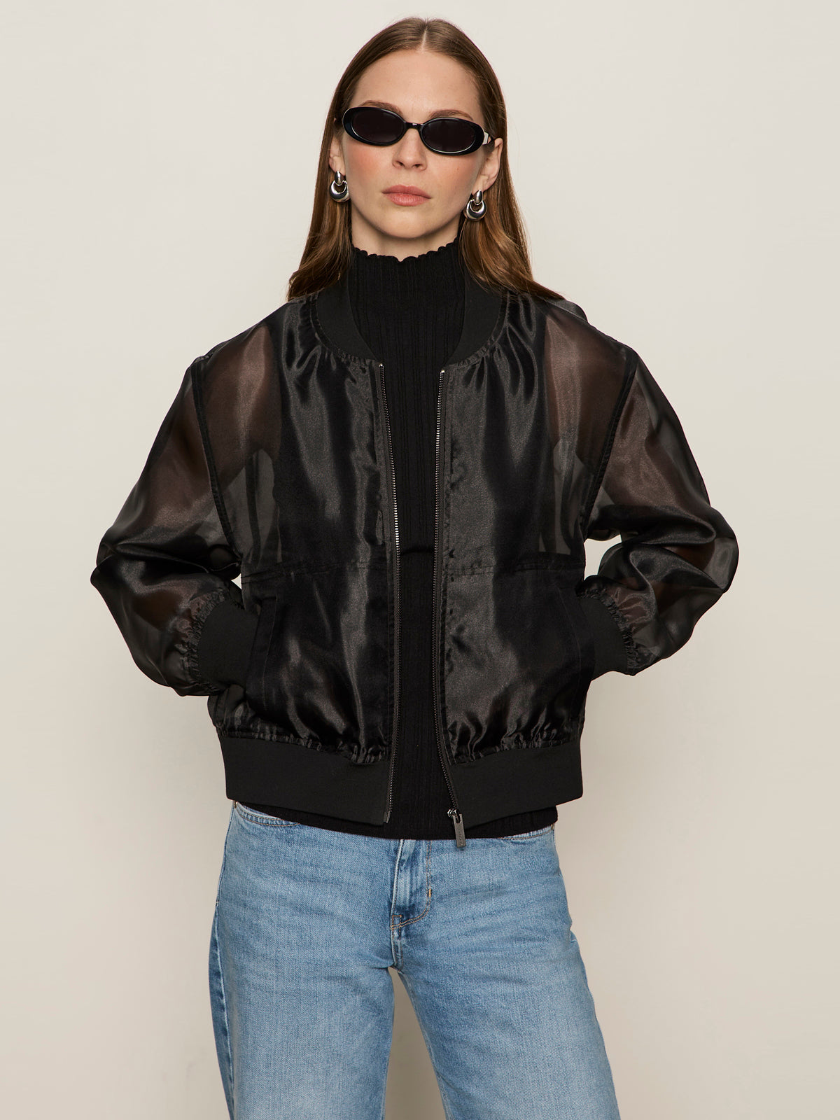 A woman wears the Sanctuary Clothing shane organza bomber black over a black top with black sunglasses and light blue jeans, standing against a plain beige background with her hands in her jacket pockets.