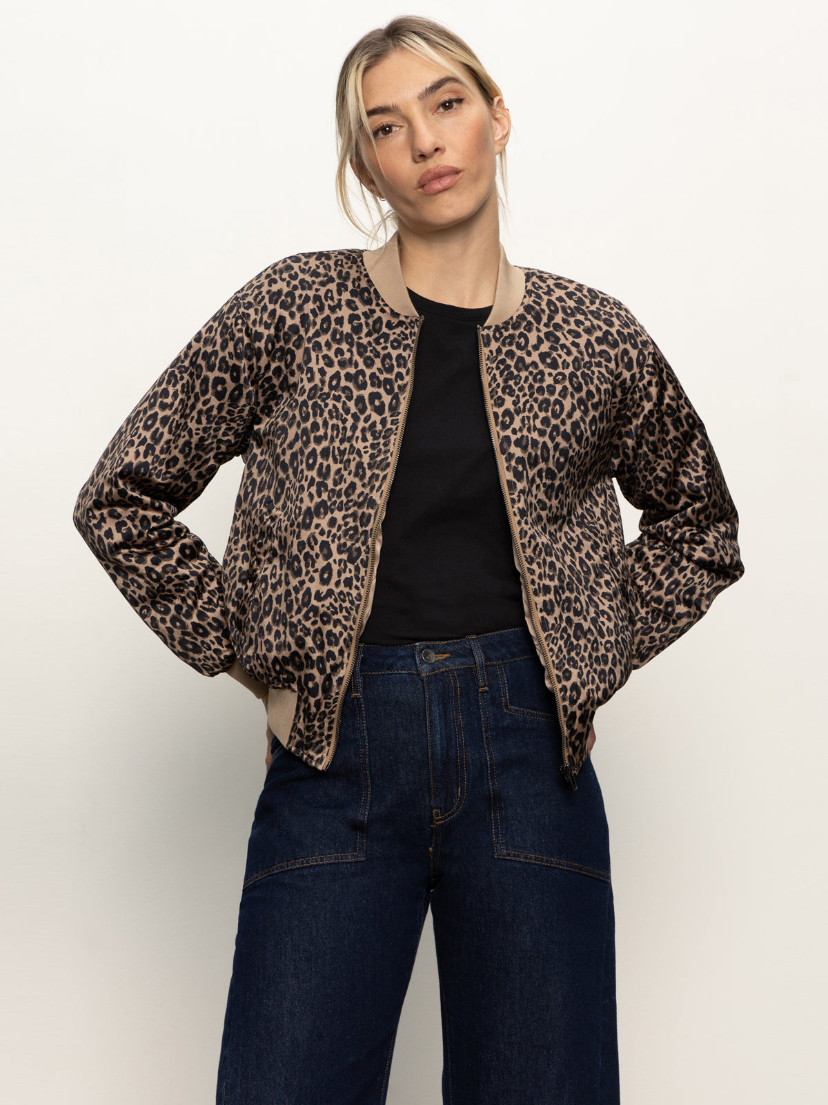 A woman wears the Sanctuary Clothing monroe sateen bomber jacket french kitty over a black shirt and dark blue jeans, standing against a plain light background with her hands in her jacket pockets.