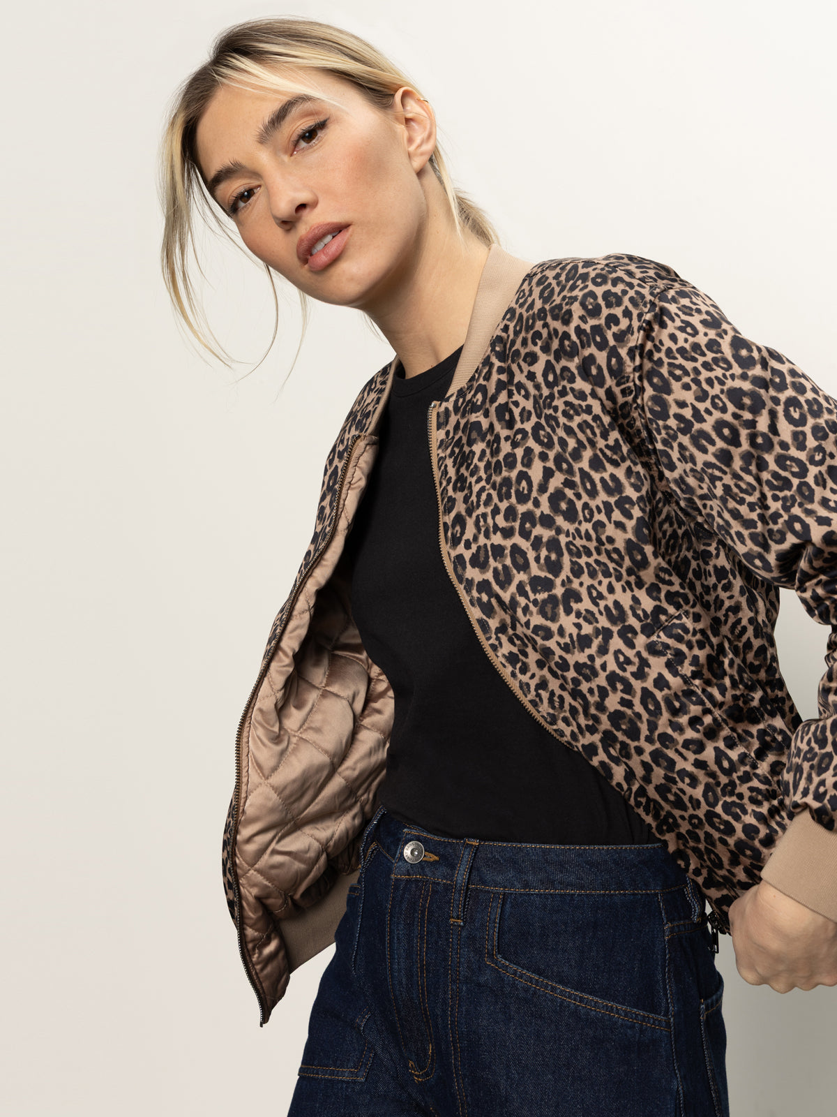 A woman with blonde hair wears the Sanctuary Clothing Monroe Sateen Bomber Jacket in French Kitty over a black top and blue jeans, standing against a plain light background and looking confidently at the camera.