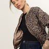 A woman with blonde hair wears the Sanctuary Clothing Monroe Sateen Bomber Jacket in French Kitty over a black top and blue jeans, standing against a plain light background and looking confidently at the camera.