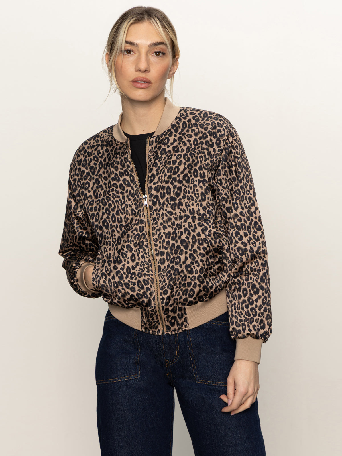 A person stands against a plain background, wearing Sanctuary Clothing’s monroe sateen bomber jacket french kitty over a black top and dark blue jeans, with their hands in the jacket pockets.
