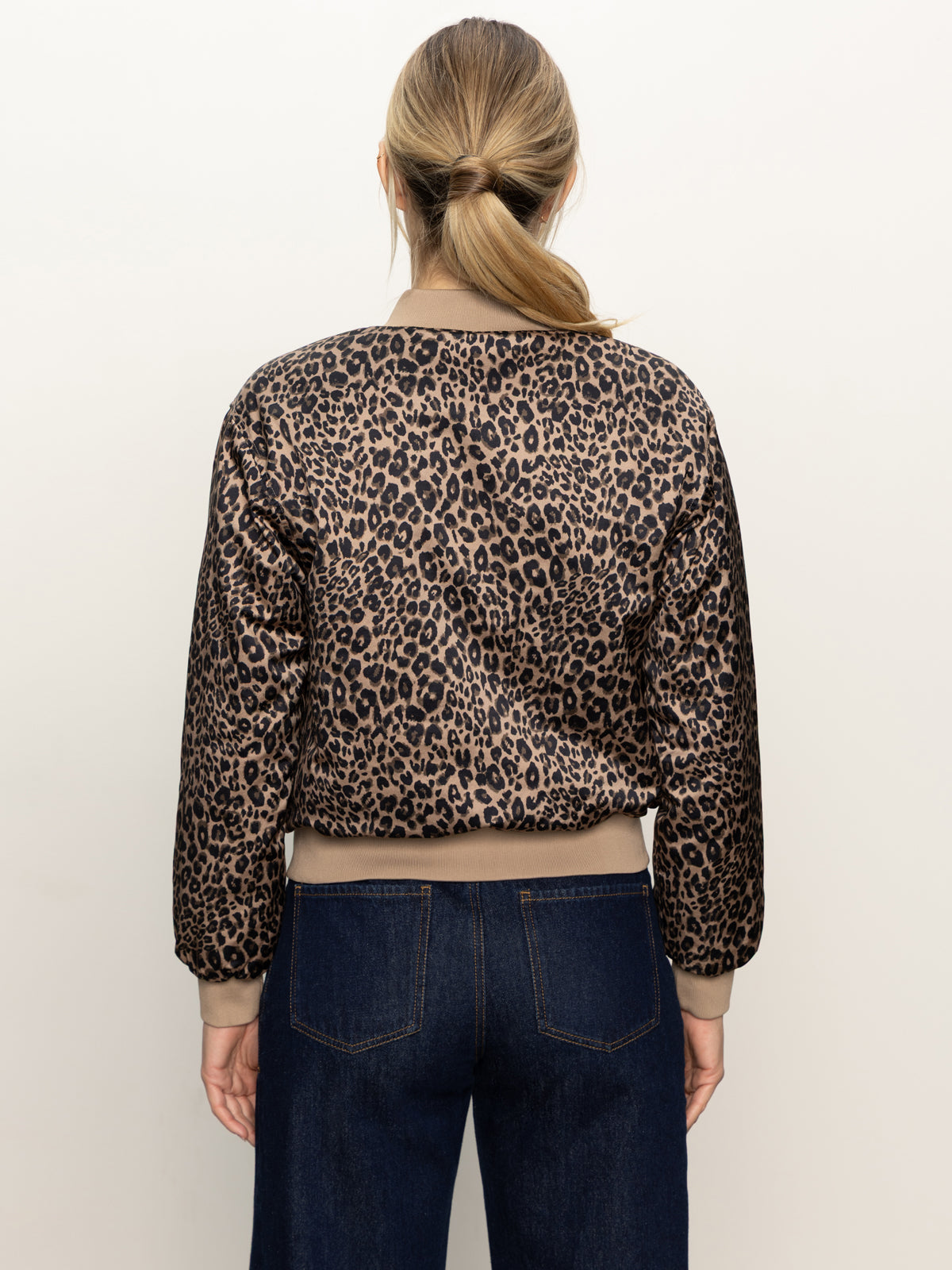 A woman with light brown hair in a low ponytail wears the Sanctuary Clothing monroe sateen bomber jacket french kitty, paired with dark blue jeans, facing away from the camera against a plain light background.