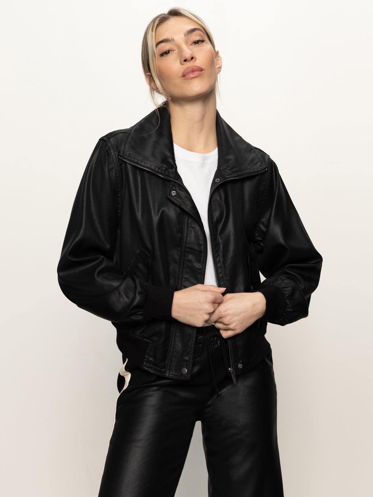 A person wearing the Sanctuary Clothing coated twill bomber jacket in black over a white shirt and black pants stands against a plain light background, looking confidently at the camera with hands in pockets.