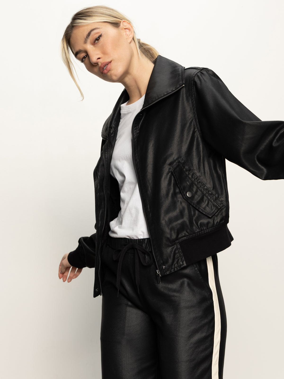 A woman models the Sanctuary Clothing coated twill bomber jacket in black over a white t-shirt and matching black pants with white side stripes, standing against a plain light background and gazing confidently at the camera.