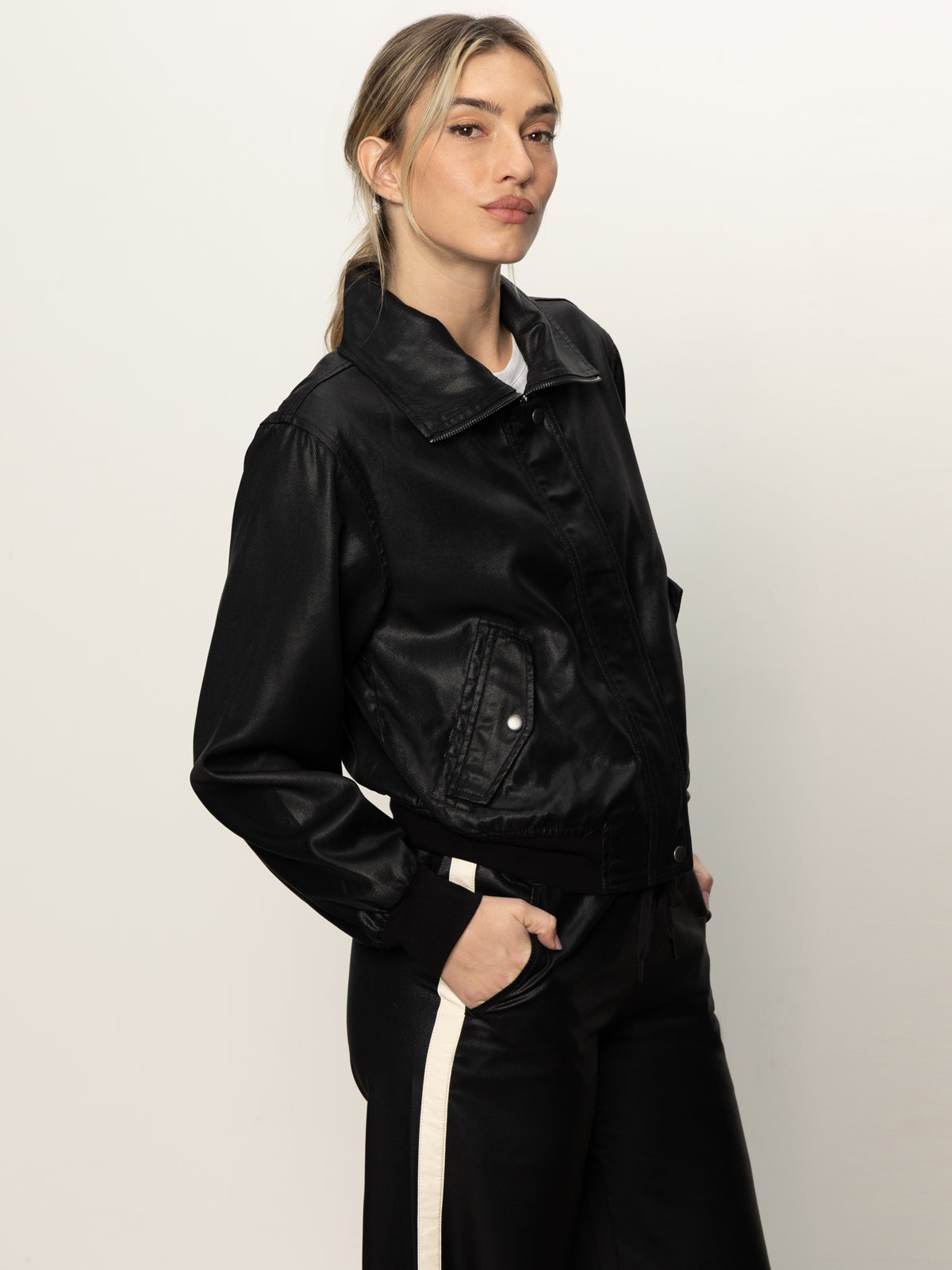 A woman with light hair tied back wears the Sanctuary Clothing coated twill bomber jacket in black and black pants with a white side stripe, standing confidently against a plain white background with one hand in her pocket.
