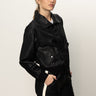 A woman with light hair tied back wears the Sanctuary Clothing coated twill bomber jacket in black and black pants with a white side stripe, standing confidently against a plain white background with one hand in her pocket.