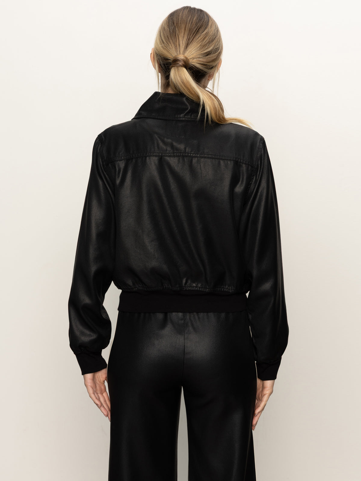 A woman with light brown hair in a low ponytail is seen from behind wearing the Sanctuary Clothing coated twill bomber jacket in black, against a plain light background.