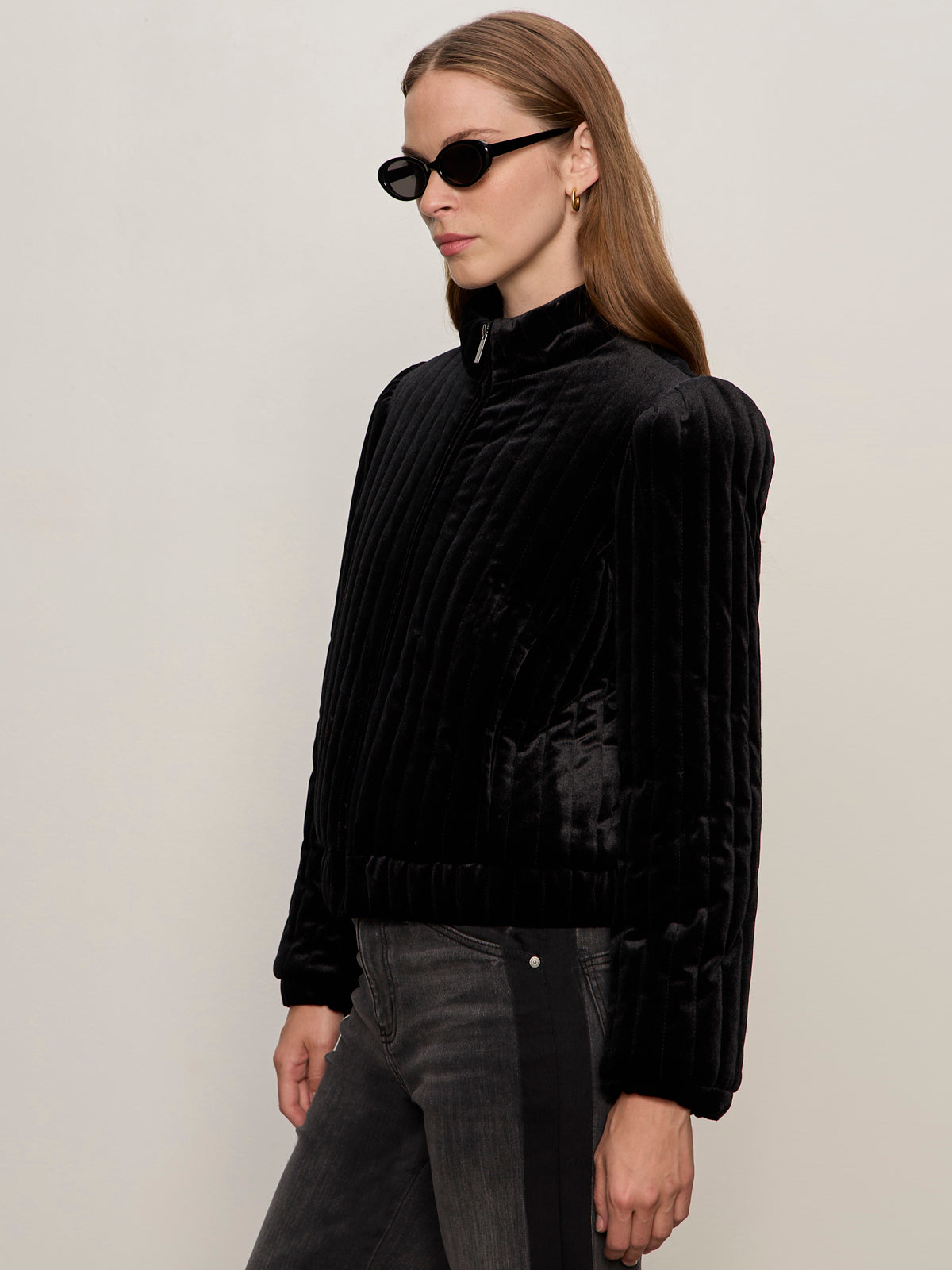 A woman with long brown hair and a neutral expression wears the Sanctuary Clothing quilted velvet jacket in black, paired with black sunglasses and dark jeans, standing against a plain light background.
