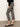A person stands sideways against a plain background wearing Sanctuary Clothing’s Carnaby Kick Crop Legging Fluid Zebra pants, paired with a black long-sleeve top and black low-heeled strap shoes.