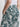A person wearing the Sanctuary Clothing "the soft trouser green oasis," featuring a high waist and tropical leaf pattern, styled with a white sleeveless top and one hand in their pocket.