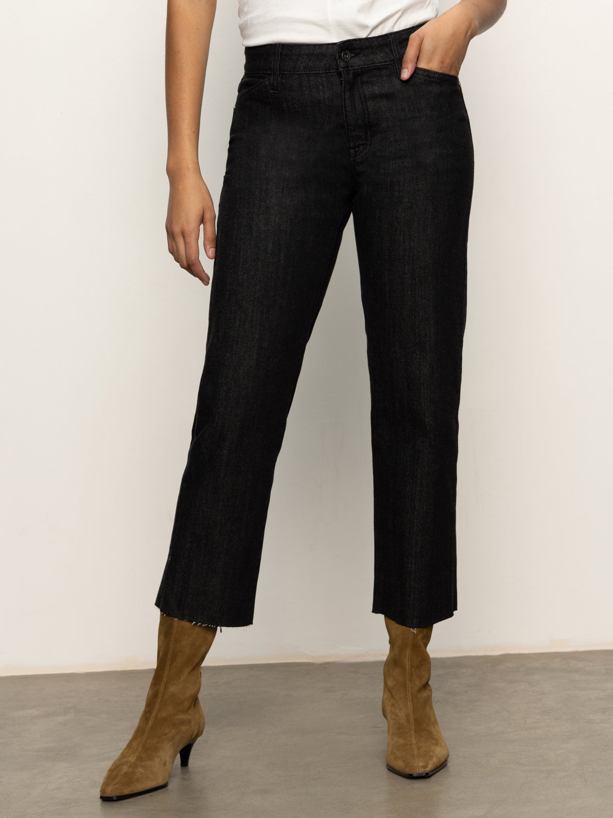 A person in Sanctuary Clothing's the marine jeans after hours—high-waisted black jeans with frayed hems—paired with a white top and tan suede ankle boots, stands against a light-colored wall.