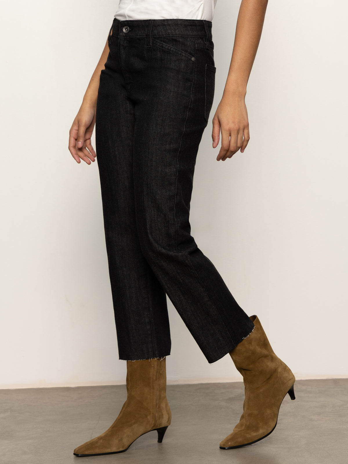 Wearing Sanctuary Clothing's "the marine jeans after hours" in black, a white top, and tan suede ankle boots with low heels, someone stands against a light wall and concrete floor.