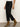 Wearing Sanctuary Clothing's "the marine jeans after hours" in black, a white top, and tan suede ankle boots with low heels, someone stands against a light wall and concrete floor.