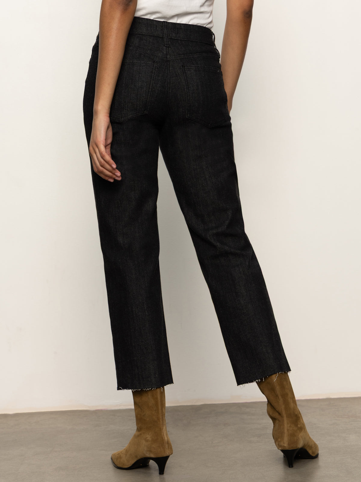 A person faces away, wearing Sanctuary Clothing's the marine jeans after hours—black high-waisted jeans with frayed hems—paired with a white top and tan suede heeled boots. The background is plain and neutral.