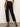 A person faces away, wearing Sanctuary Clothing's the marine jeans after hours—black high-waisted jeans with frayed hems—paired with a white top and tan suede heeled boots. The background is plain and neutral.