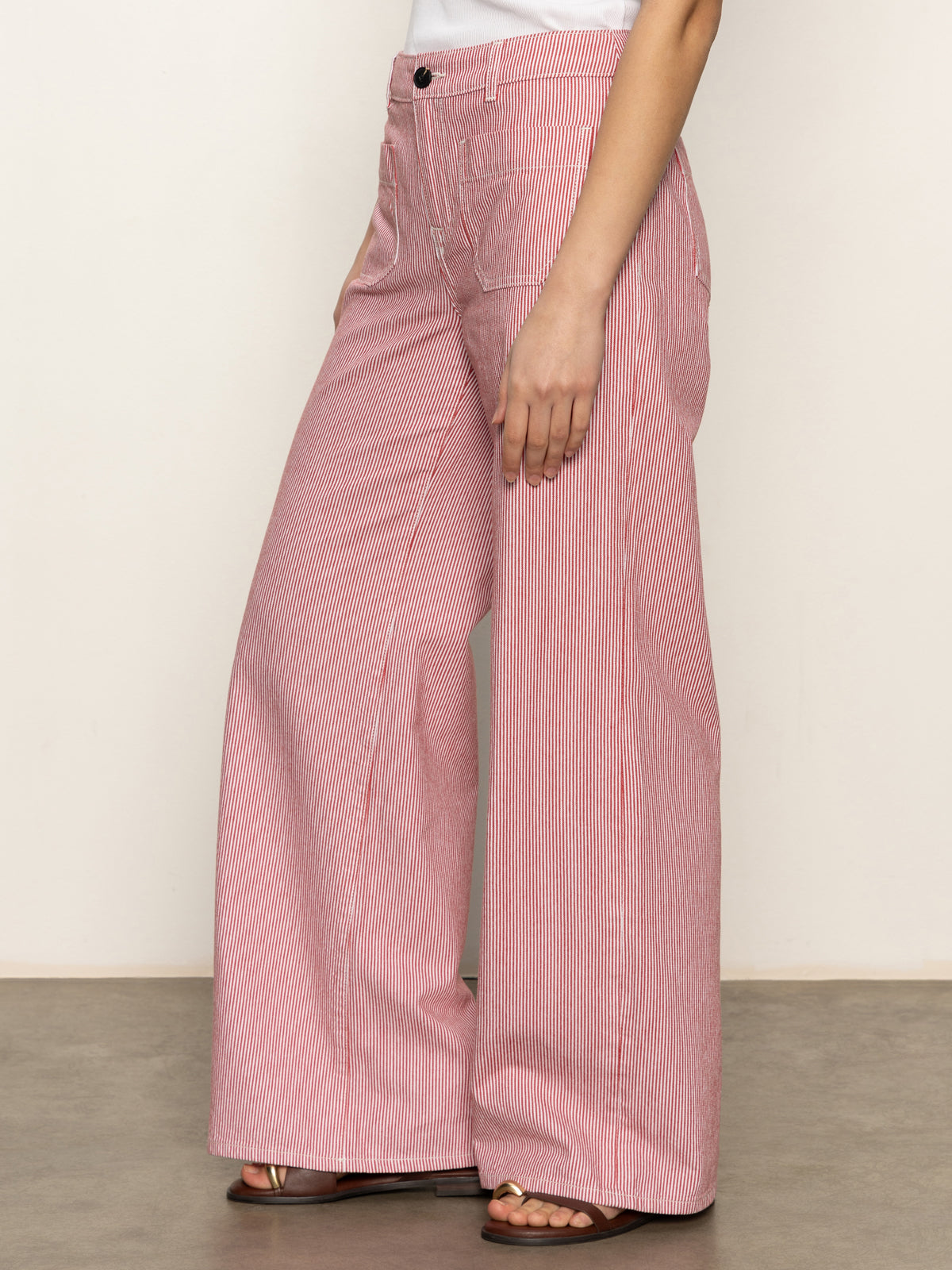 A person in Sanctuary Clothing's marine full length semi high rise pant galley stripe stands sideways with hands in pockets, wearing sandals and a white top against a plain neutral background.