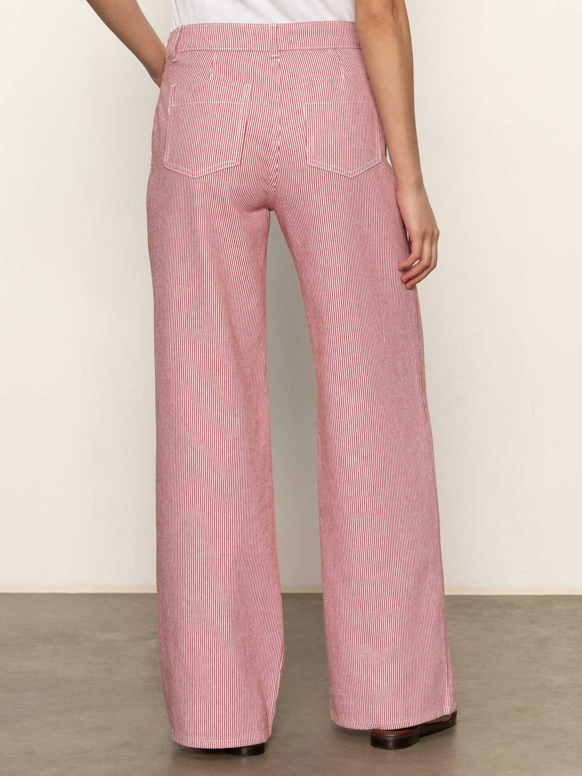 A person faces away from the camera, wearing Sanctuary Clothing's marine full length semi high rise pant galley stripe—red and white striped, wide-leg pants with back pockets—and a white top, standing on a smooth, light-colored floor.