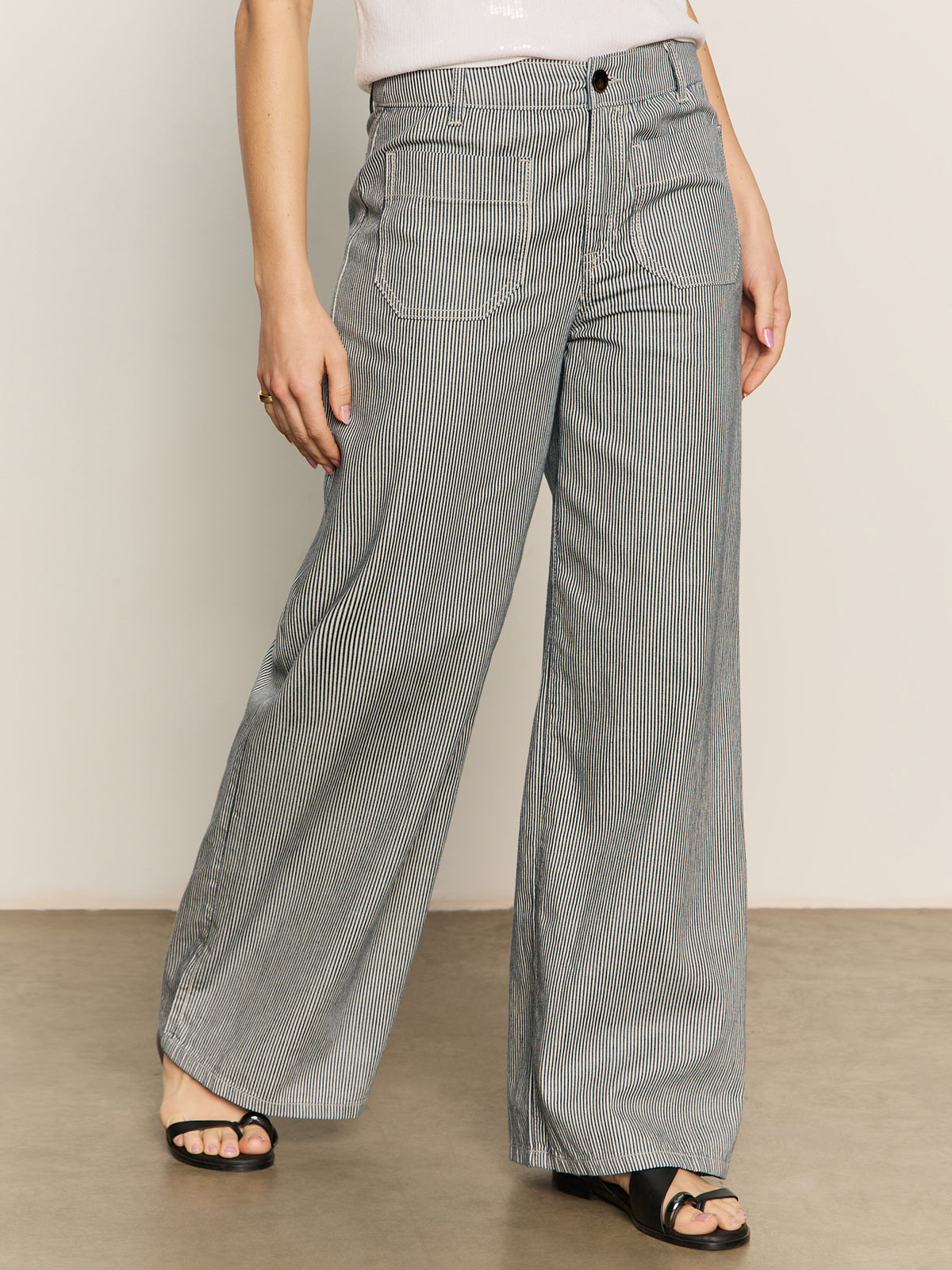 A person stands against a plain background wearing Sanctuary Clothing’s marine full length semi high rise pant in yacht stripe. The gray pinstriped pants are paired with black open-toe sandals; the photo is cropped from the waist down.