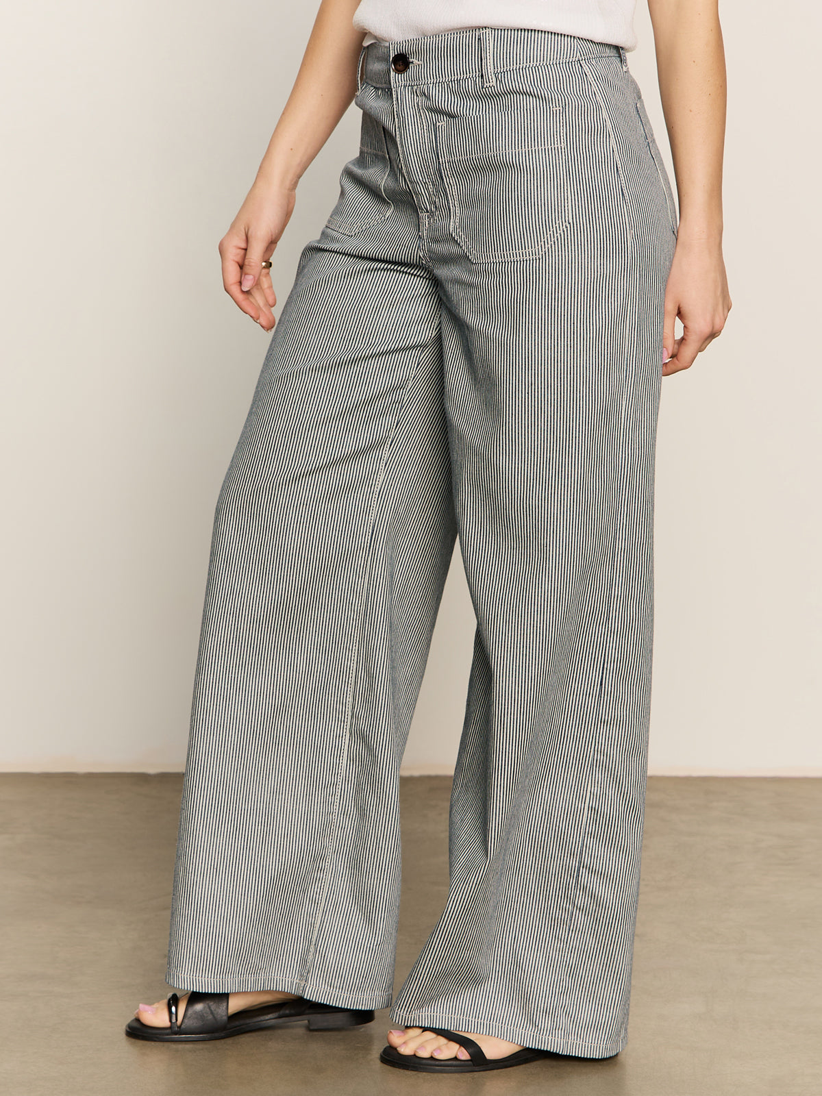 A person wears Sanctuary Clothing's marine full length semi high rise pant yacht stripe with black sandals and a light top, standing on a smooth, light-colored floor against a plain background.