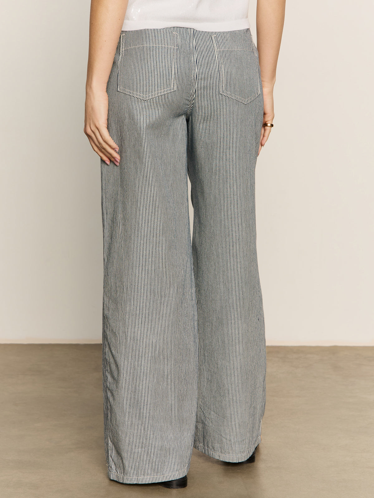 A person stands on a light floor, shown from the back, wearing Sanctuary Clothing’s marine full length semi high rise pant in yacht stripe—wide-leg pants with thin black and white vertical stripes, two back pockets, and a white top.