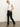A woman with blonde hair in a ponytail wears Sanctuary Clothing's sculpted hayden bootcut black jeans, a white sleeveless top, and tan heeled boots. She stands on a gray floor against a white background, posing with one hand in her pocket.