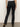 A person faces away, wearing Sanctuary Clothingâ‚¬„¢s Sculpted Hayden Bootcut Black pants with flap pockets on the back and legs, paired with a white top and brown heeled shoes against a plain light background.