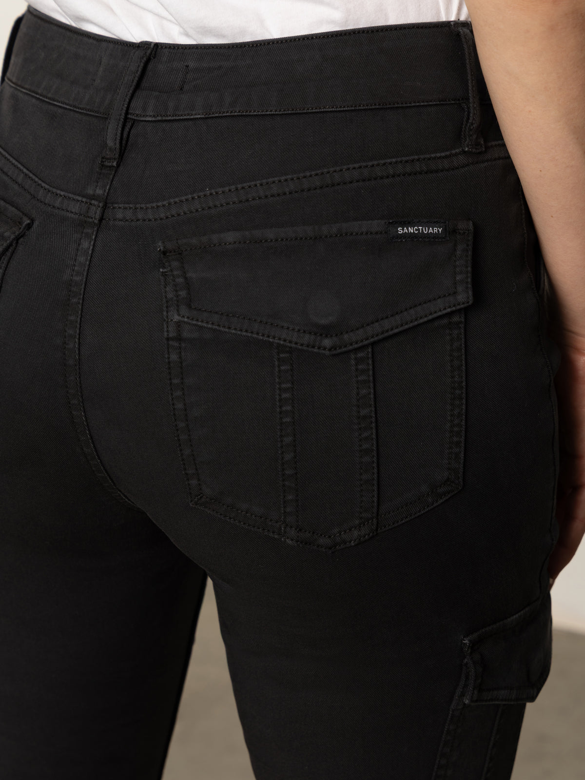 Close-up of a person wearing Sanctuary Clothing's sculpted hayden bootcut black jeans, featuring a flap back pocket with a small black "Sanctuary" label. A white shirt is tucked in.