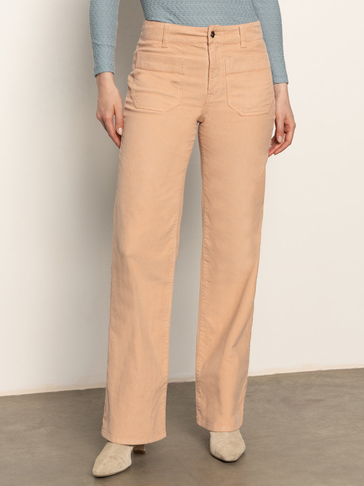 A person stands against a plain wall, wearing Sanctuary Clothing's corduroy marine pant in dusty rose, paired with a light blue long-sleeve top and beige shoes; their head is out of frame.