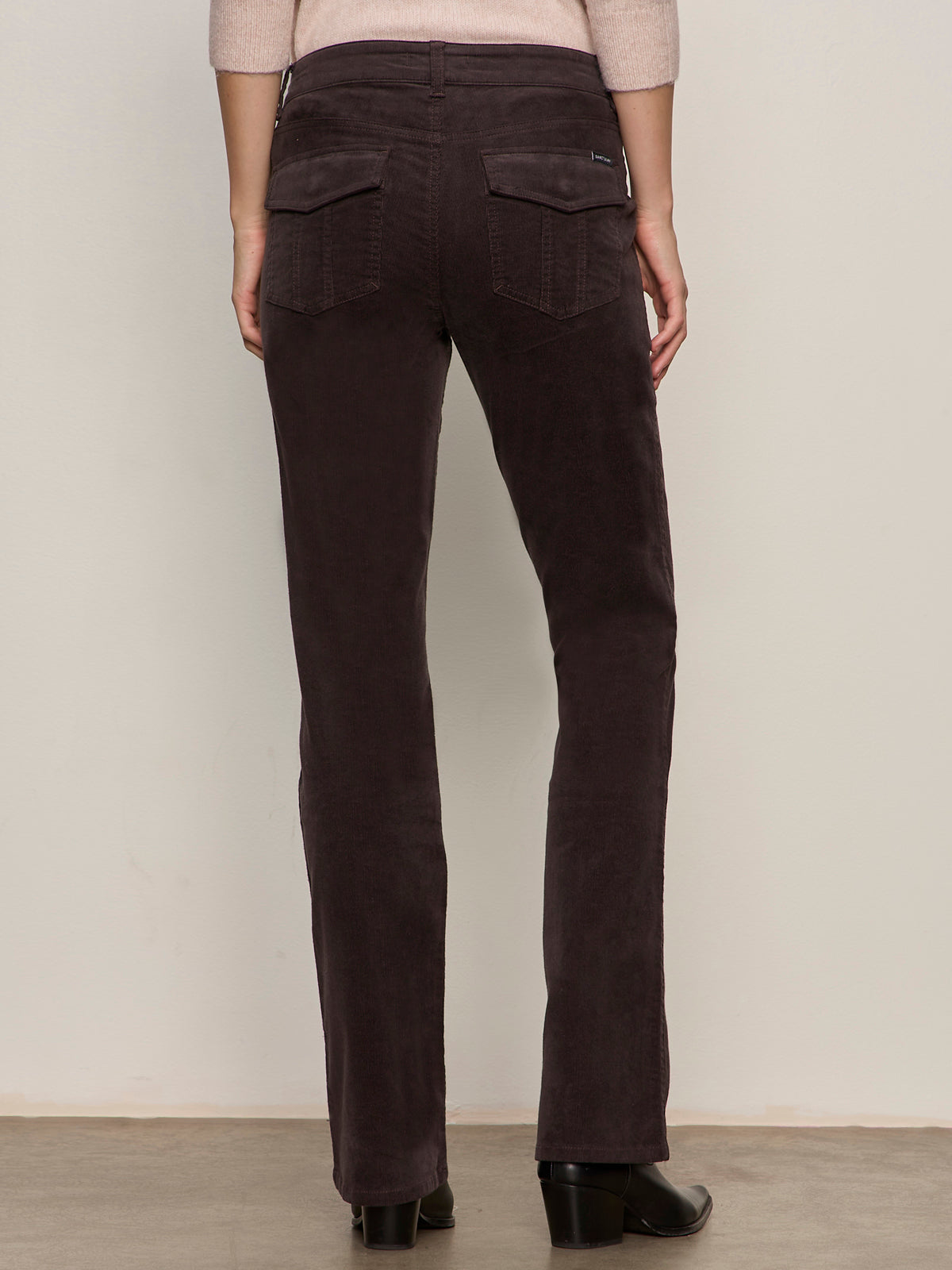 A person shown from the back wears Sanctuary Clothing’s Corduroy Hayden Bootcut Pant in Semi Sweet, featuring two flap back pockets, paired with black shoes and standing against a light wall.
