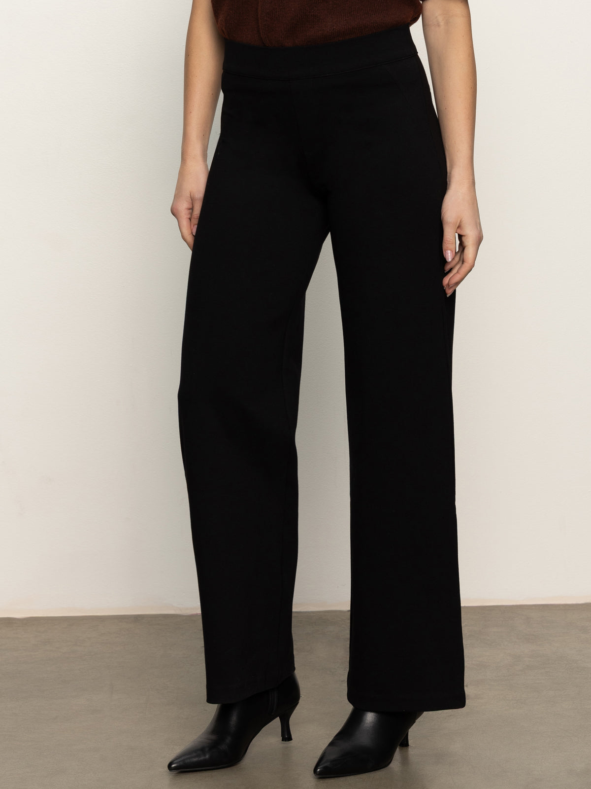 A person wears the Sanctuary Clothing runway ponte trouser in black, paired with black heeled ankle boots, standing against a plain light-colored wall and brown floor. The top half of their body is partially visible.