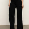 A person wears the Sanctuary Clothing runway ponte trouser in black, paired with black heeled ankle boots, standing against a plain light-colored wall and brown floor. The top half of their body is partially visible.