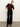 A woman stands against a plain white wall wearing a brown short-sleeve sweater, black heeled boots, and the Sanctuary Clothing runway ponte trouser in black, with her hands tucked behind her back and her head slightly tilted.