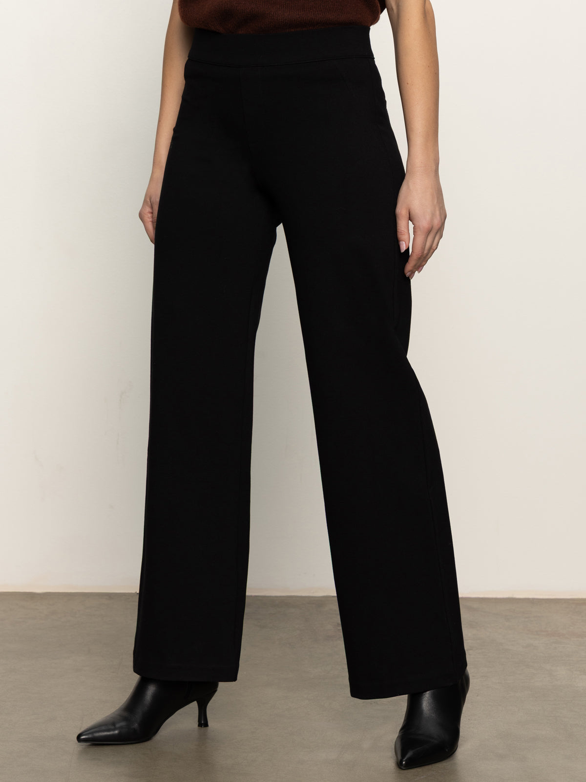 Wearing the Sanctuary Clothing runway ponte trouser in black with black heeled ankle boots, standing on a gray floor against an off-white wall; only the lower body is visible.