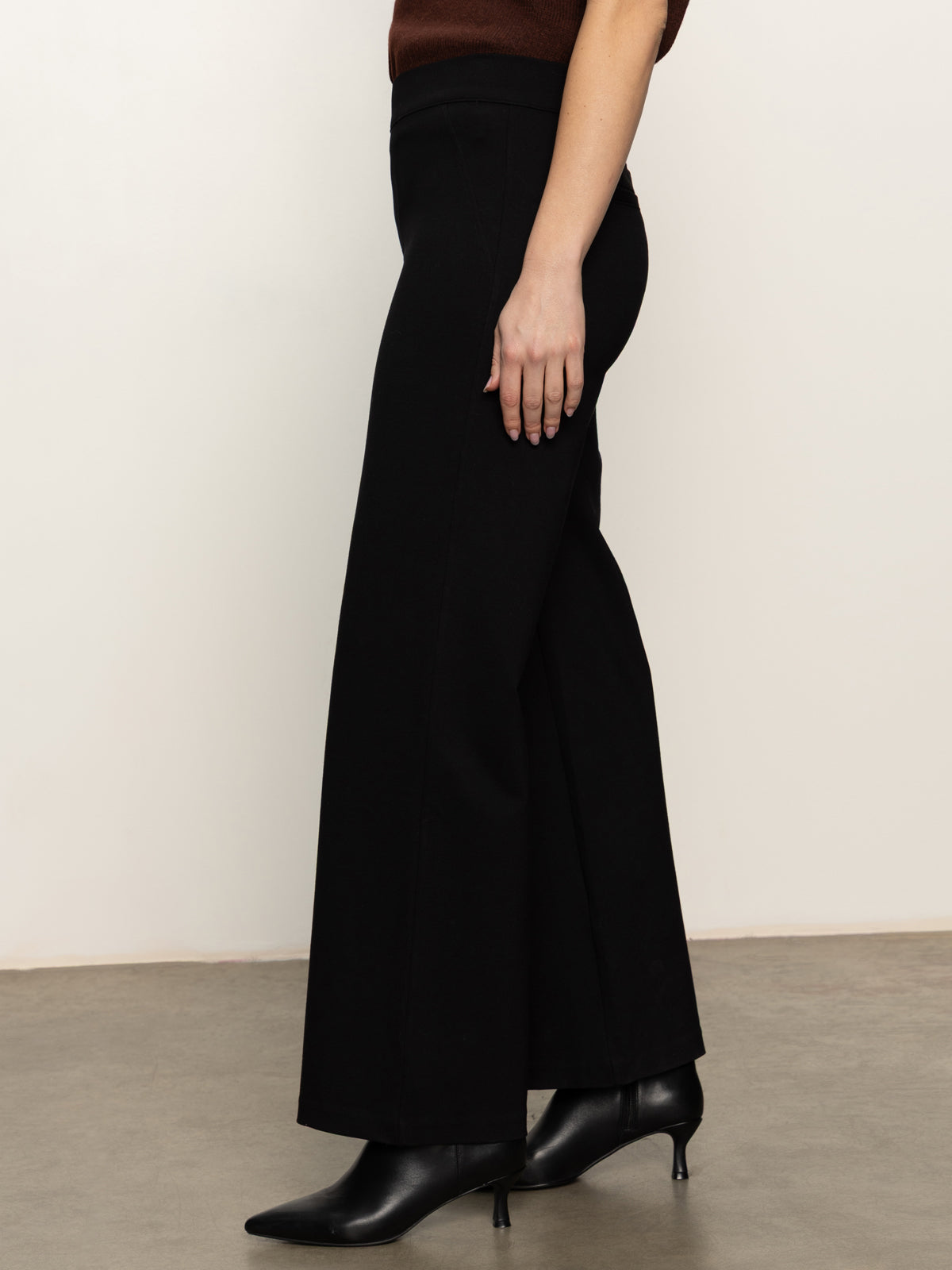A person wears the Sanctuary Clothing runway ponte trouser in black, paired with pointed black ankle boots. Only their lower half is shown, standing on a gray floor against a plain light-colored wall.