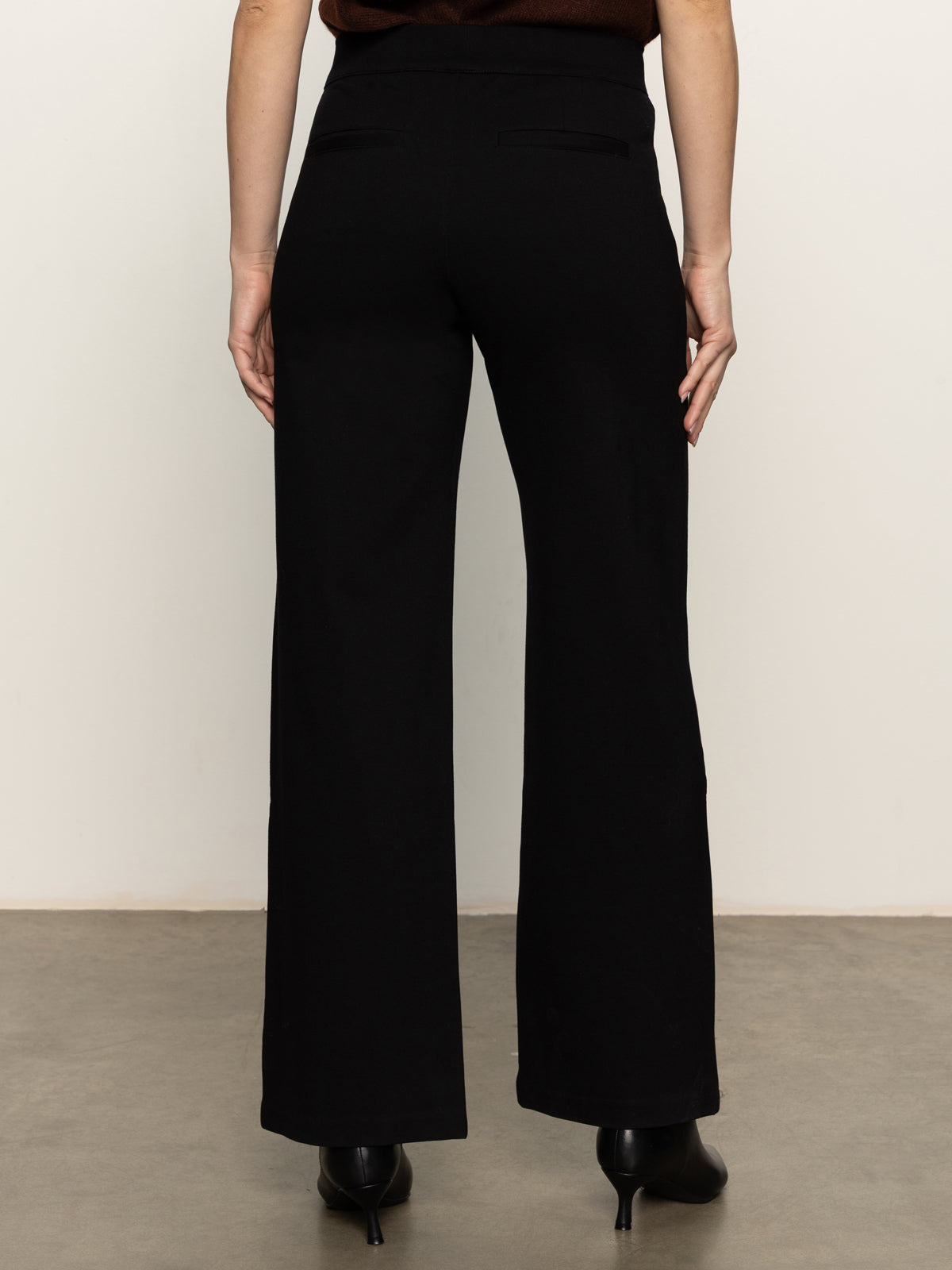 A person is seen from behind, showcasing Sanctuary Clothing's runway ponte trouser in black with heeled shoes against a neutral backdrop—only their lower back, legs, and part of the arms are visible.
