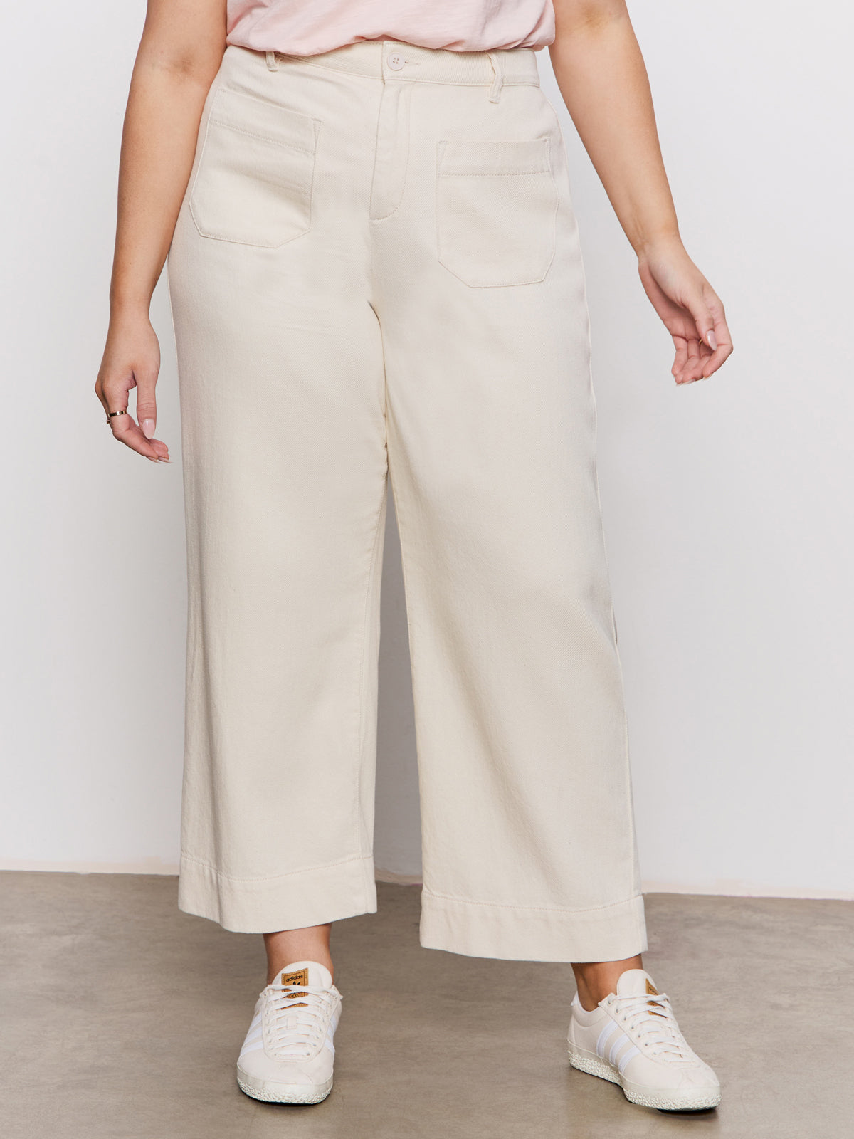 A person wears Sanctuary Clothing's soft marine wide leg crop pant macrame in cream, paired with a pink top and white sneakers, standing on a concrete floor against a plain white wall.