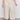 A person wears Sanctuary Clothing's soft marine wide leg crop pant macrame in cream, paired with a pink top and white sneakers, standing on a concrete floor against a plain white wall.